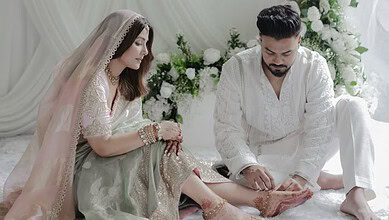 A woman and man in traditional white attire sitting on a white bed with floral decorations.