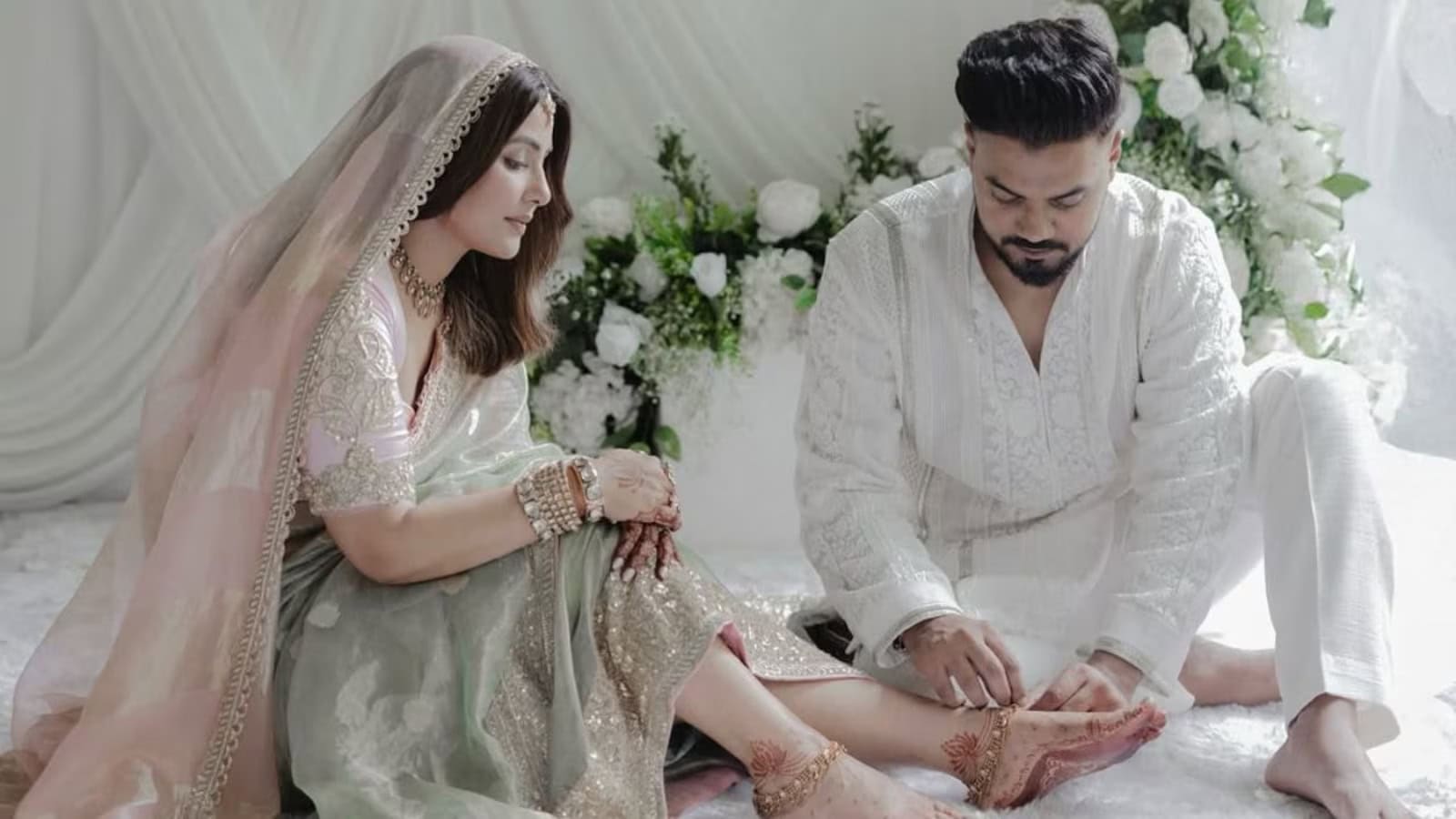 A woman and man in traditional white attire sitting on a white bed with floral decorations.