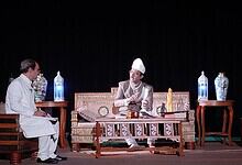 Historical scene with Salar Jung III in traditional attire on stage with two men, ornate vases, and stage.
