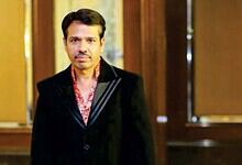 Indian man in formal black suit with red patterned shirt, standing indoors with wooden decor background.