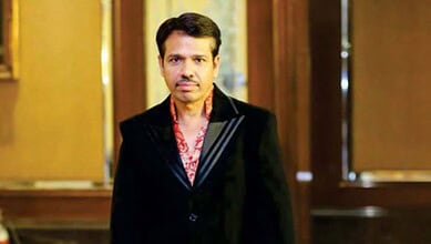 Indian man in formal black suit with red patterned shirt, standing indoors with wooden decor background.