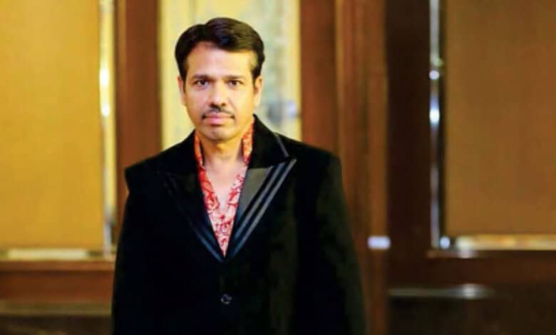Indian man in formal black suit with red patterned shirt, standing indoors with wooden decor background.
