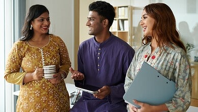 A woman in traditional yellow attire holding a mug, a man in a purple kurta with a tablet, and a woman wi.