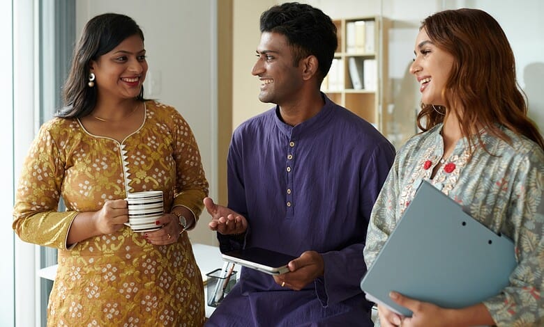 A woman in traditional yellow attire holding a mug, a man in a purple kurta with a tablet, and a woman wi.