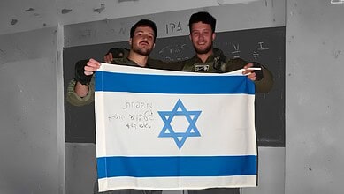 Two IDF soldiers holding an Israeli flag in a classroom setting.