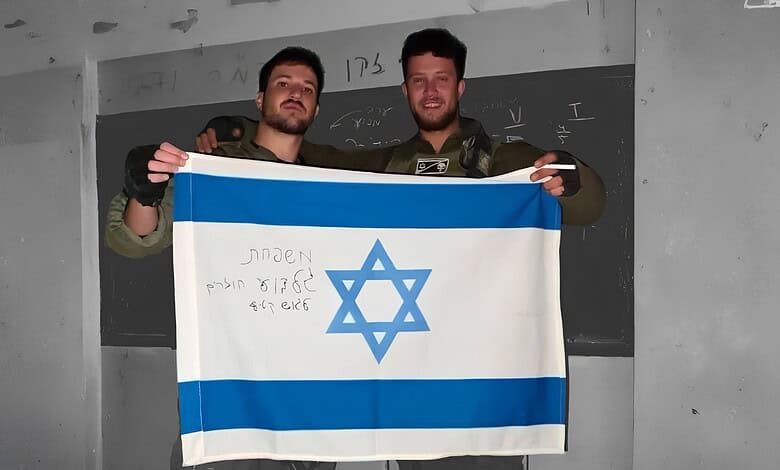 Two IDF soldiers holding an Israeli flag in a classroom setting.