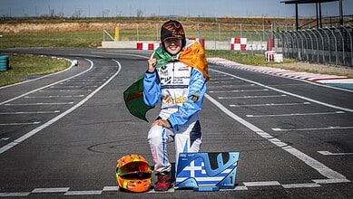 Young race car driver celebrating victory on the track with a medal and flag, wearing racing gear and hel.