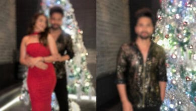 Bollywood actor in black and gold shirt with woman in red dress near Christmas trees.