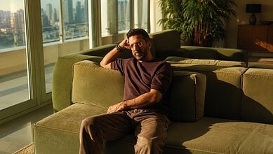Jaaved Jaaferi sitting on a green sofa in a bright, modern living room with city views.