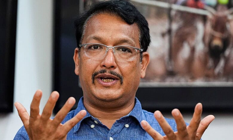 A man speaking passionately during a discussion or interview, wearing glasses and a blue shirt.