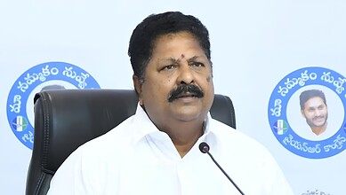 A woman in a white shirt speaking at a press conference with a microphone, seated in front of a blue logo.
