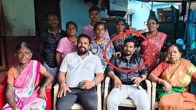 A woman in a black saree and a man in a white shirt sitting in the centre surrounded by a group of women.