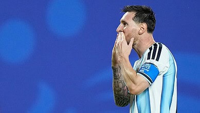 Lionel Messi in Argentina jersey, celebrating with hand over mouth, blue background.