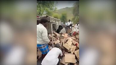 Group of people sorting and stacking cardboard boxes outdoors in a rural setting with trees and hills in.