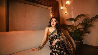 Elegant woman wearing a black and gold saree seated on a beige sofa at The Siasat Daily event, with warm.