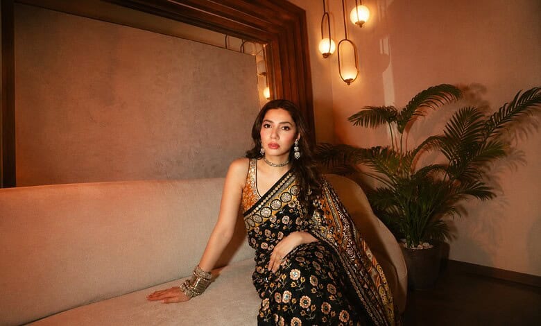 Elegant woman wearing a black and gold saree seated on a beige sofa at The Siasat Daily event, with warm.