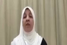 A woman wearing a white hijab and black attire speaking passionately during a public event.