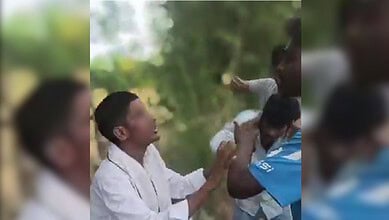 A woman in white holding a child outdoors, with another person in a blue shirt nearby, in a natural green.