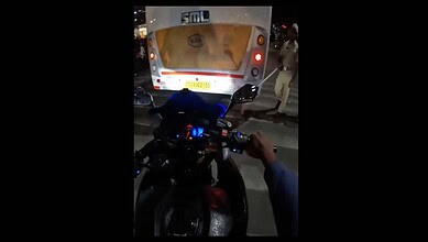 A person riding a motorcycle at night with a bus in front, illuminated by streetlights, in an urban setti.