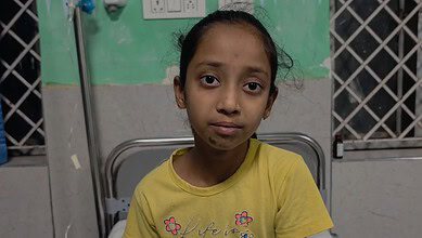 Young girl with a serious expression in a hospital room, wearing a yellow T-shirt.
