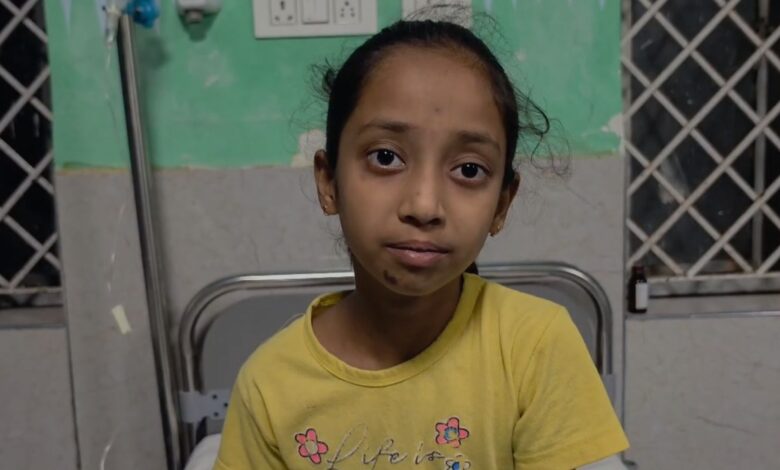 Young girl with a serious expression in a hospital room, wearing a yellow T-shirt.