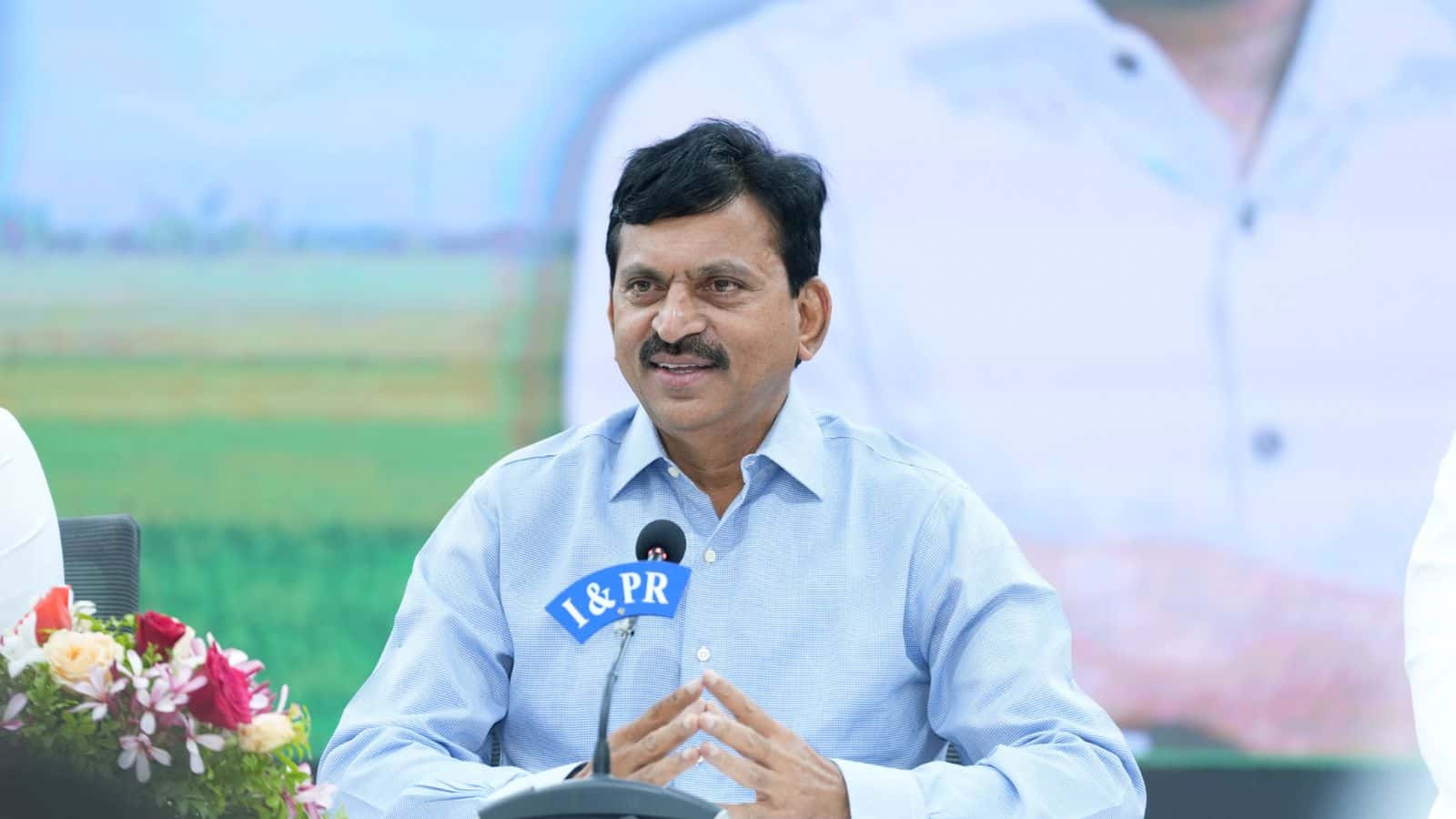 A man in a light blue shirt speaking at a press conference outdoors with a microphone labeled