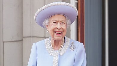 Queen Elizabeth II smiling in a light blue dress and hat, standing outdoors with a building in the背景.