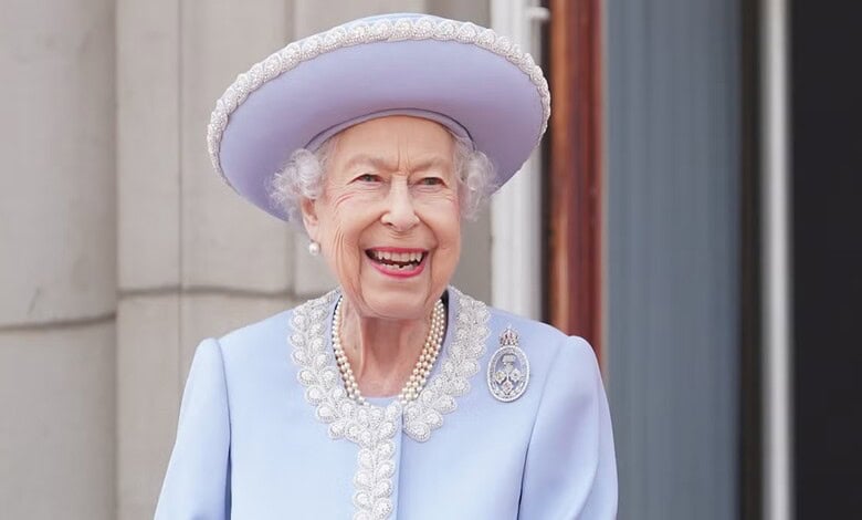 Queen Elizabeth II smiling in a light blue dress and hat, standing outdoors with a building in the背景.