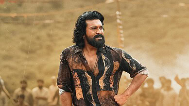 A man with long hair and a beard stands confidently outdoors, wearing a patterned shirt, with a blurred c.