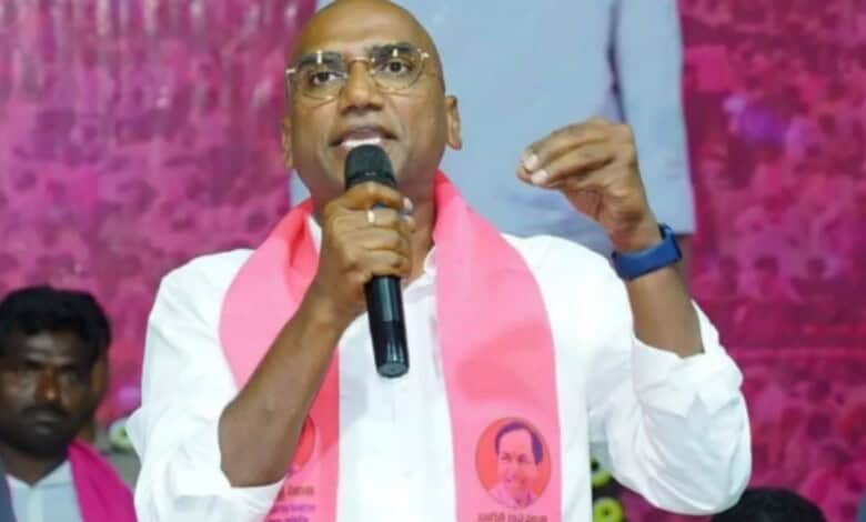 Prominent man speaking passionately at a public event, wearing a white shirt and pink scarf, with a pink.