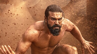 A man with a beard and intense expression, covered in dirt, in a dynamic action pose amidst a dusty backg.
