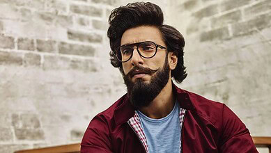 A man with glasses and a beard, wearing a maroon jacket and blue shirt, looking thoughtfully against a br.