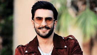 Smiling man with sunglasses and beard wearing a brown jacket outdoors.
