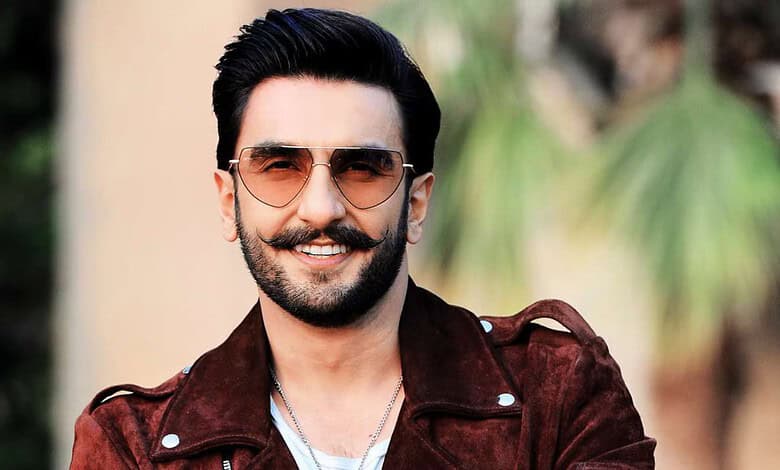Smiling man with sunglasses and beard wearing a brown jacket outdoors.
