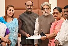A woman in pink saree and glasses presenting a document to a senior man with grey hair and beard, surroun.