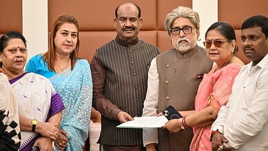 A woman in pink saree and glasses presenting a document to a senior man with grey hair and beard, surroun.