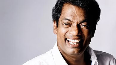 A smiling man with dark curly hair wearing a white shirt against a light grey background.