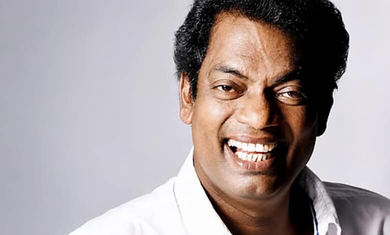 A smiling man with dark curly hair wearing a white shirt against a light grey background.