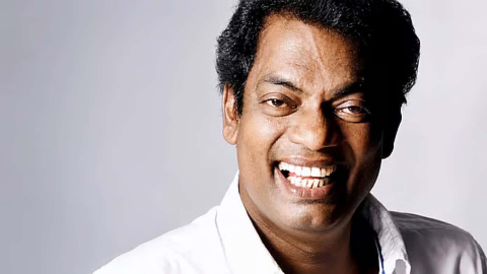 A smiling man with dark curly hair wearing a white shirt against a light grey background.