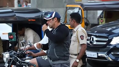 Salman Khan in casual attire on a cycle at a film set, surrounded by vehicles and crew members.