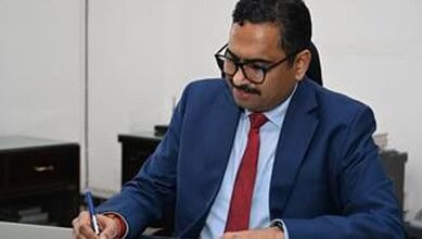 Sanjay Jaju in a formal suit, sitting at a desk, looking down, in an office setting.
