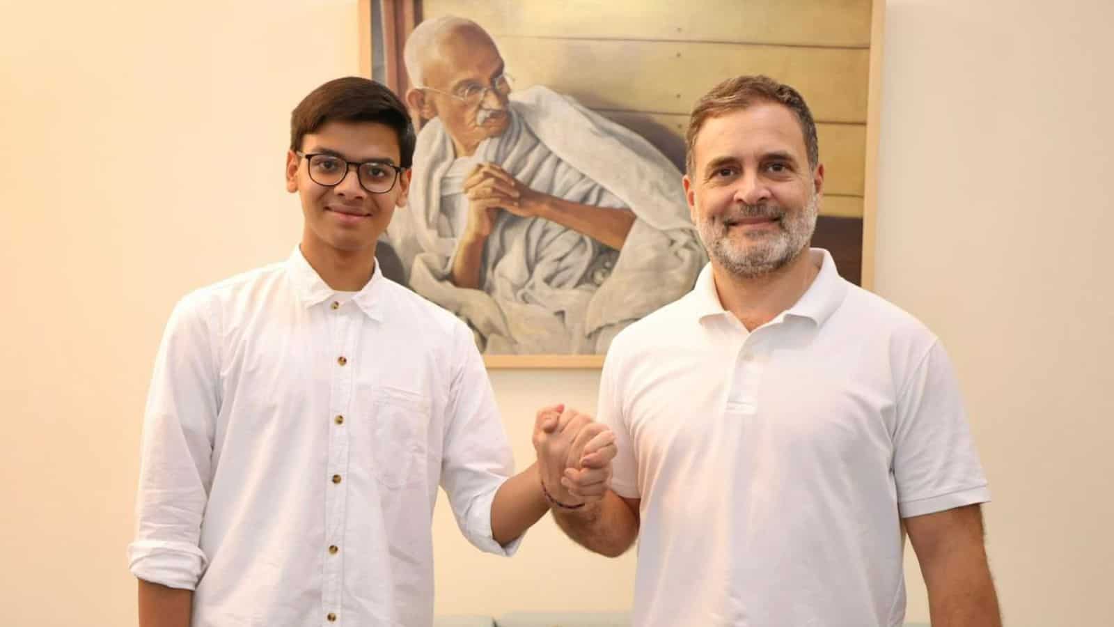 A young man and an older man shaking hands in front of a portrait of Mahatma Gandhi.