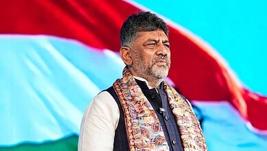 Indian politician standing with eyes closed during a patriotic event, with the Indian flag waving behind.
