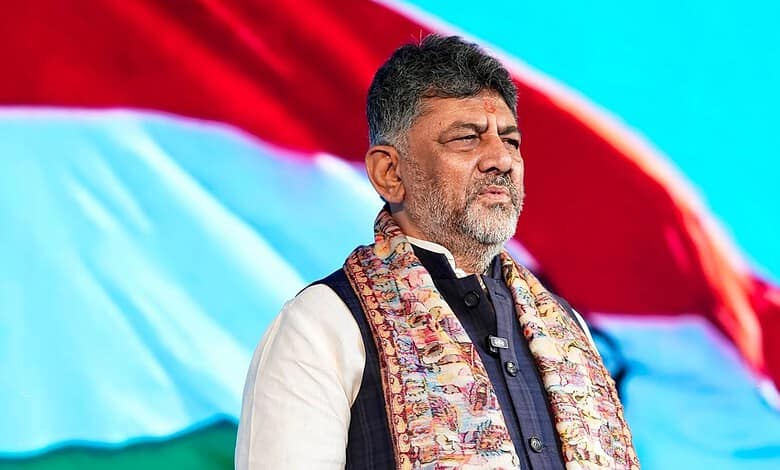 Indian politician standing with eyes closed during a patriotic event, with the Indian flag waving behind.