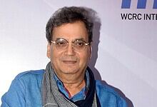 A middle-aged Indian man with glasses, wearing a blue shirt and scarf, attending a formal event with a ba.