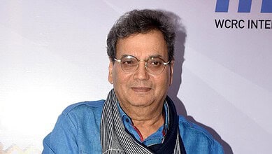A middle-aged Indian man with glasses, wearing a blue shirt and scarf, attending a formal event with a ba.