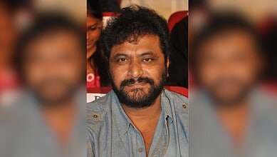 A man with curly black hair and a beard, wearing a denim shirt, sitting in a red auditorium seat at an ev.