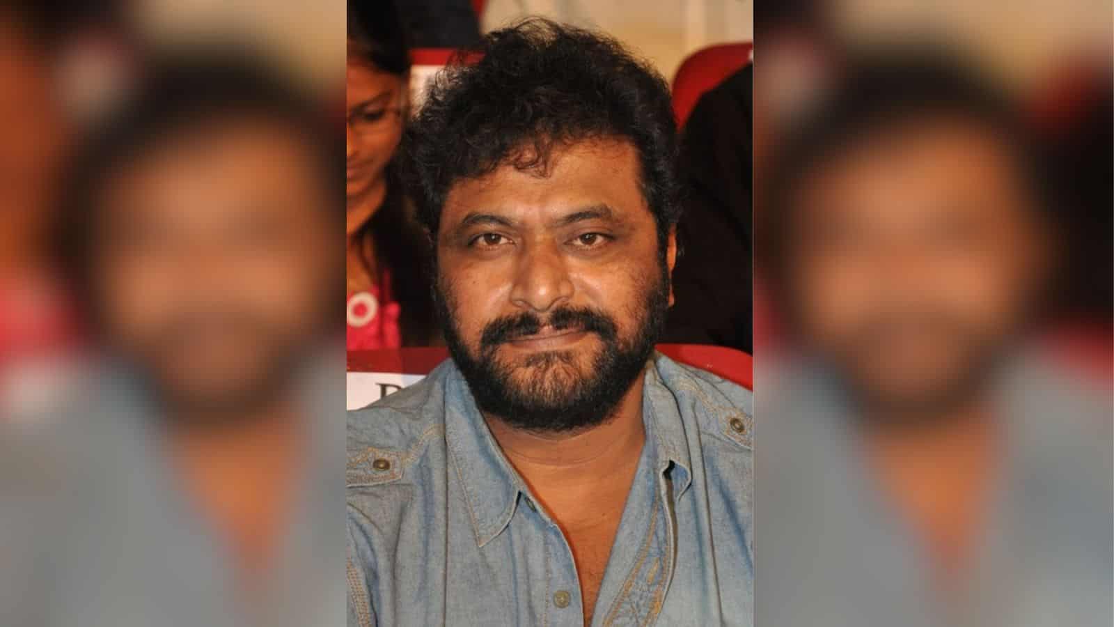 A man with curly black hair and a beard, wearing a denim shirt, sitting in a red auditorium seat at an ev.