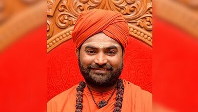 A man dressed in traditional orange attire with a turban, smiling, seated on an ornate wooden chair with.