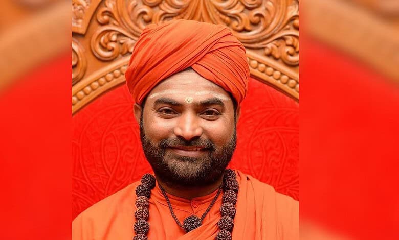 A man dressed in traditional orange attire with a turban, smiling, seated on an ornate wooden chair with.